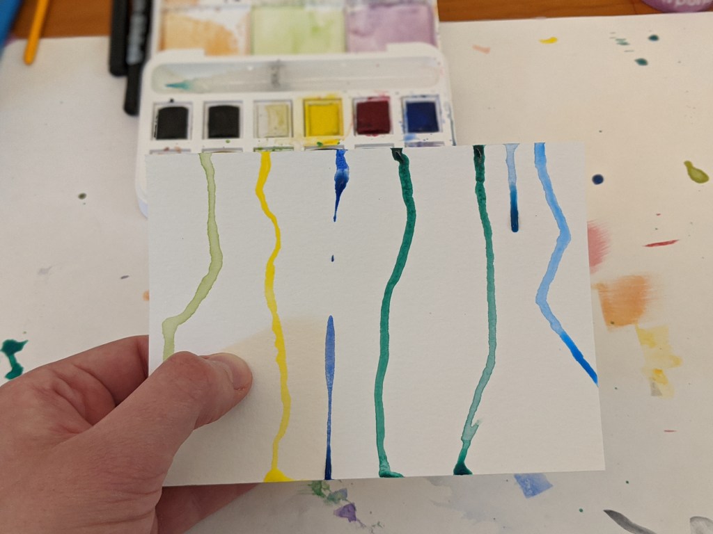 Heather's hand is holding a postcard-sized piece of paper with green, yellow, and blue drops of wet watercolor paint running down it.
