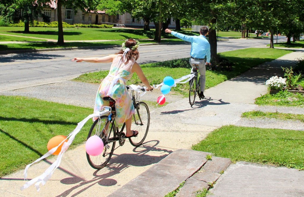 Two figures bike away from the camera, each waving their left arm goodbye. The first figure is wearing a multicolored dress and the second a nice shirt and pants. Their bikes have streamers and balloons, for they were just married.