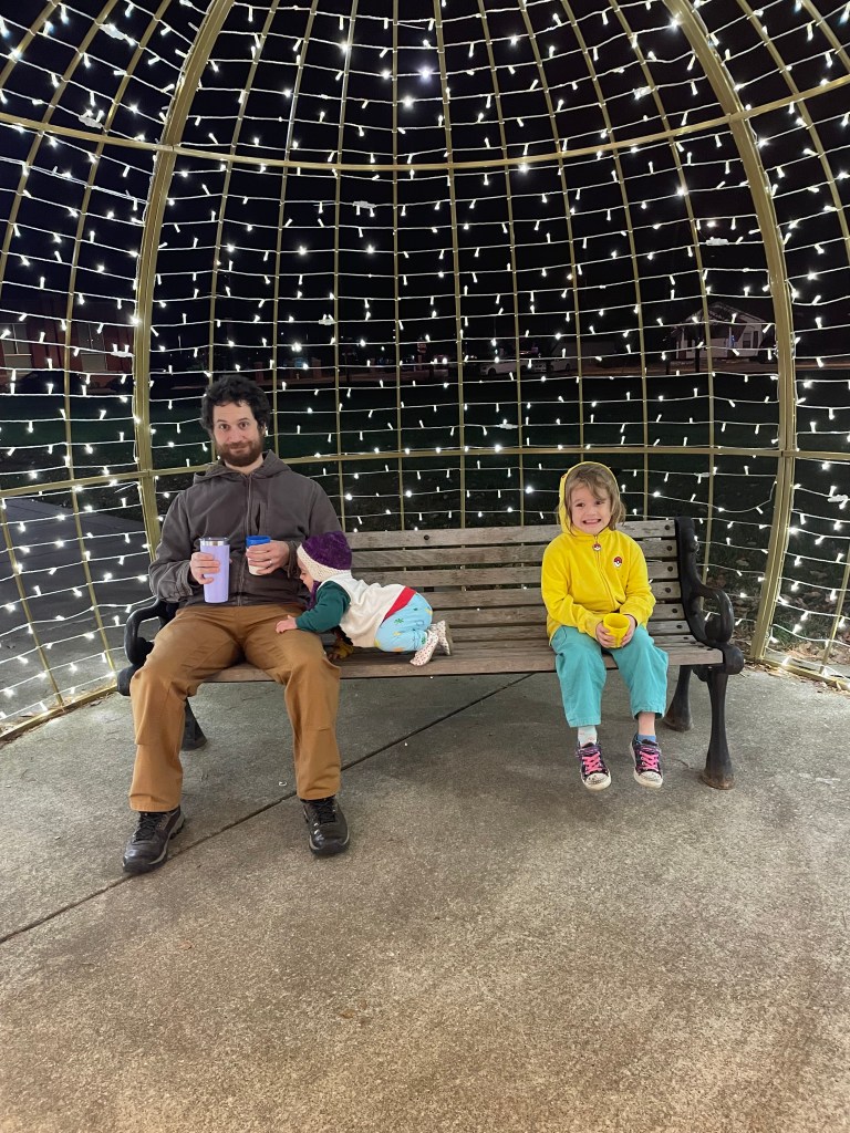 Three people, one big, two small, sit in on a bench under a lights display made round like an ornament. The baby isn't looking at the camera.