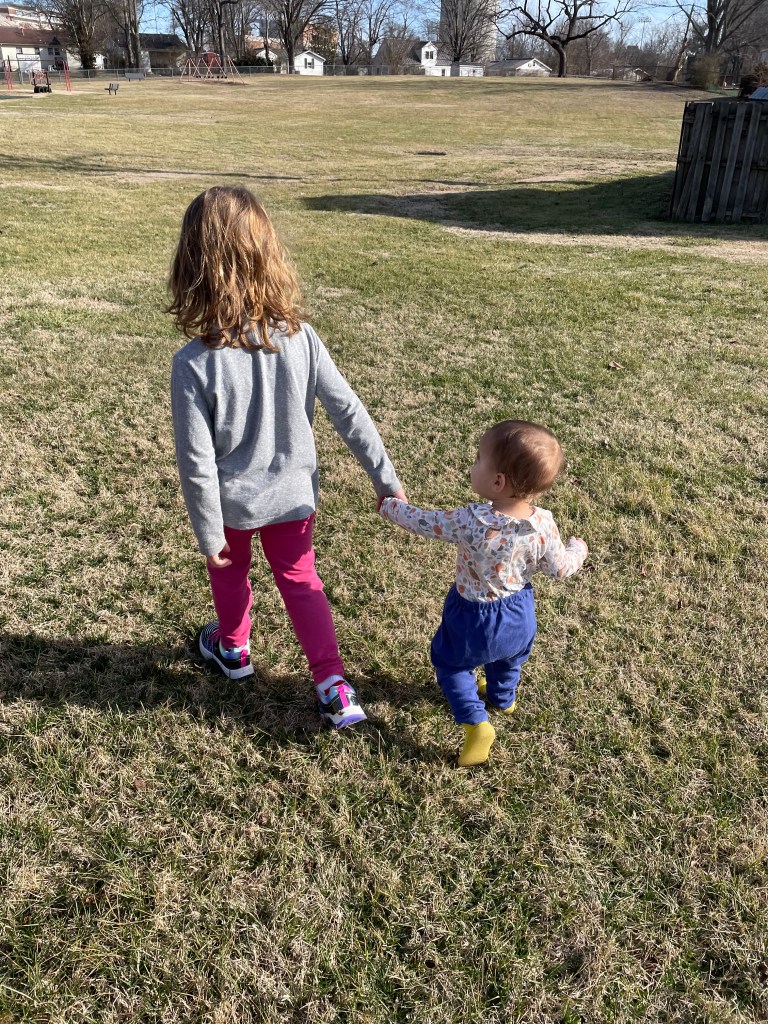 Two kids, one toddler and one a kindergartener, walk hand-in-hand in an open field on a bright, sunny day.
