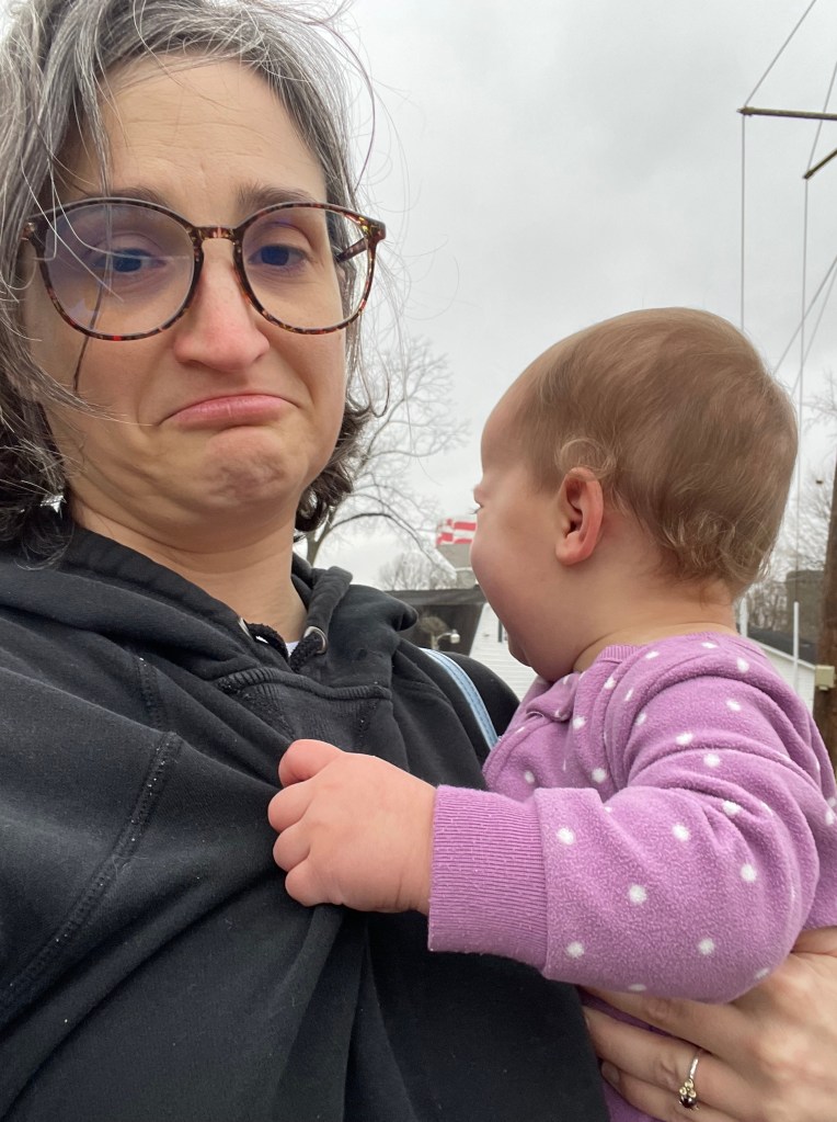 Mom holding a crying young toddler makes a sad face at the camera. Alas, they are not going to the playground today.
