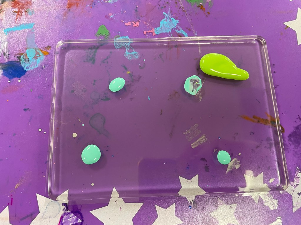 A clear rectangular gel has five dollops of paint, four teal and one light green