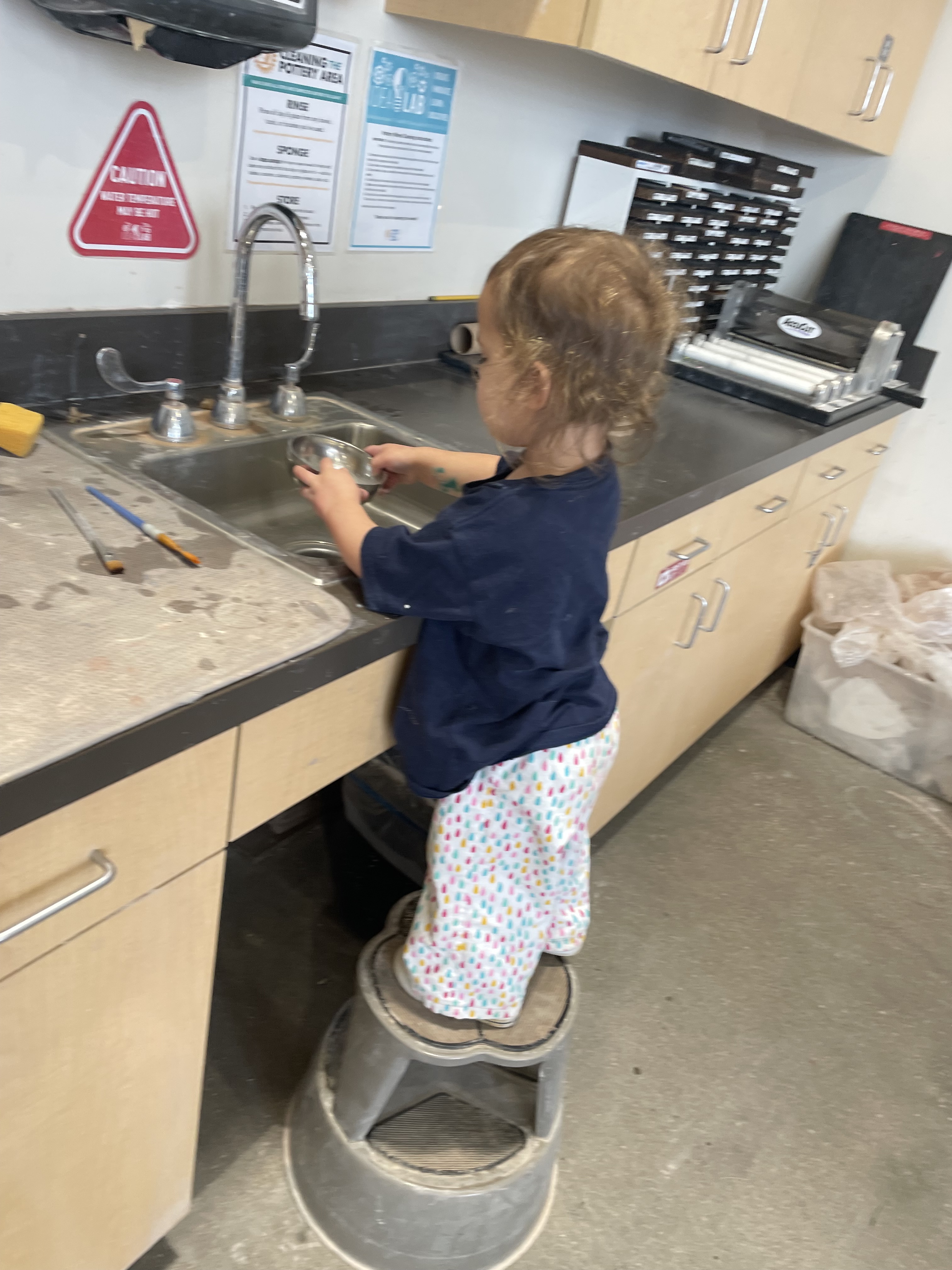 Juniper washes brushes while standing on a library step stool. 