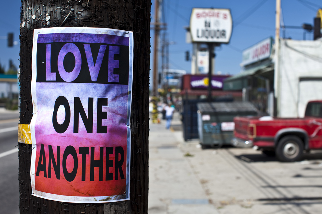 In the foreground, a flyer attached to a telephone poll says "Love One Another". In the background, we see a pickup truck and a liquor store.